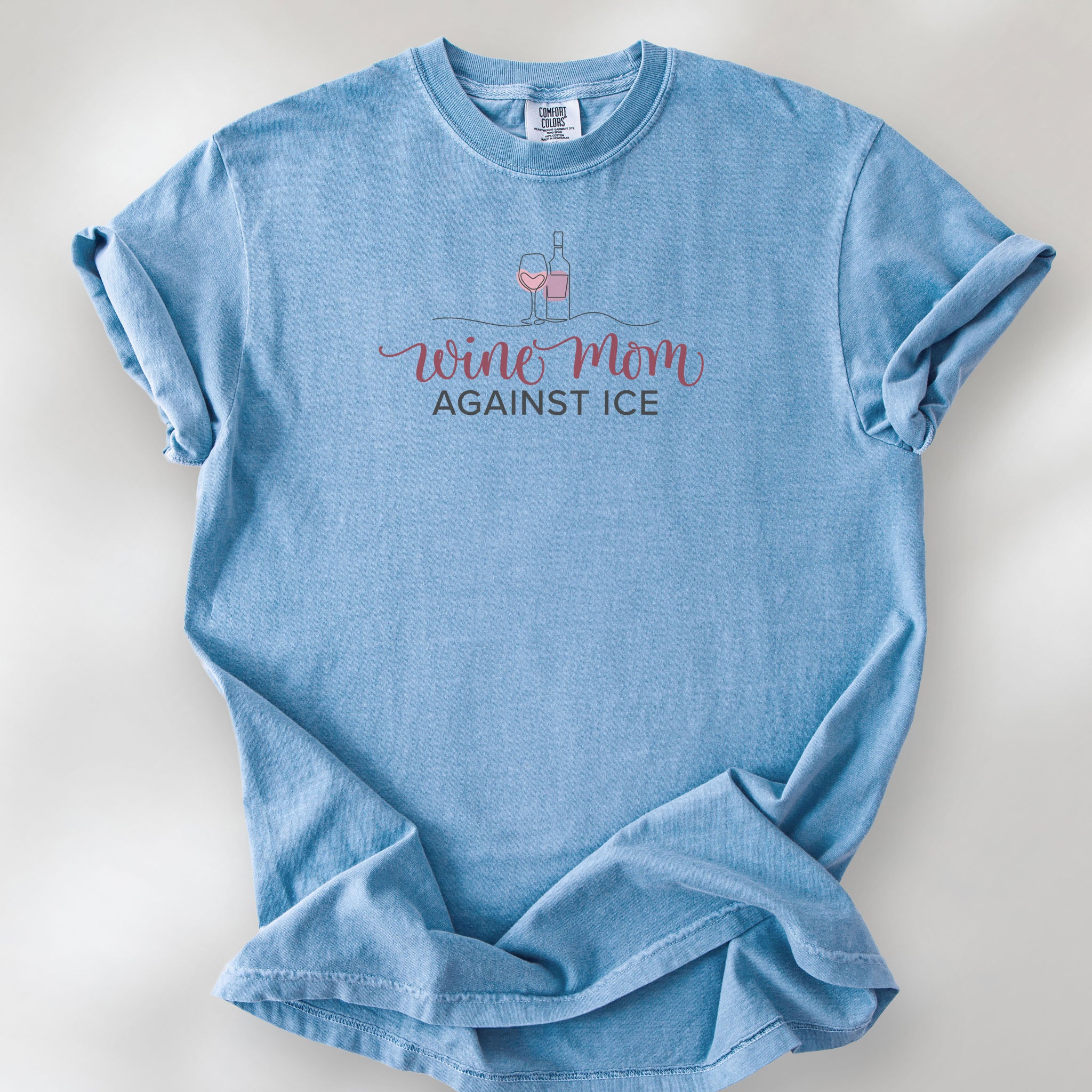 Washed denim t-shirt featuring the text “Wine Mom Against ICE” with a minimalist illustration of a wine bottle and glass above the lettering.