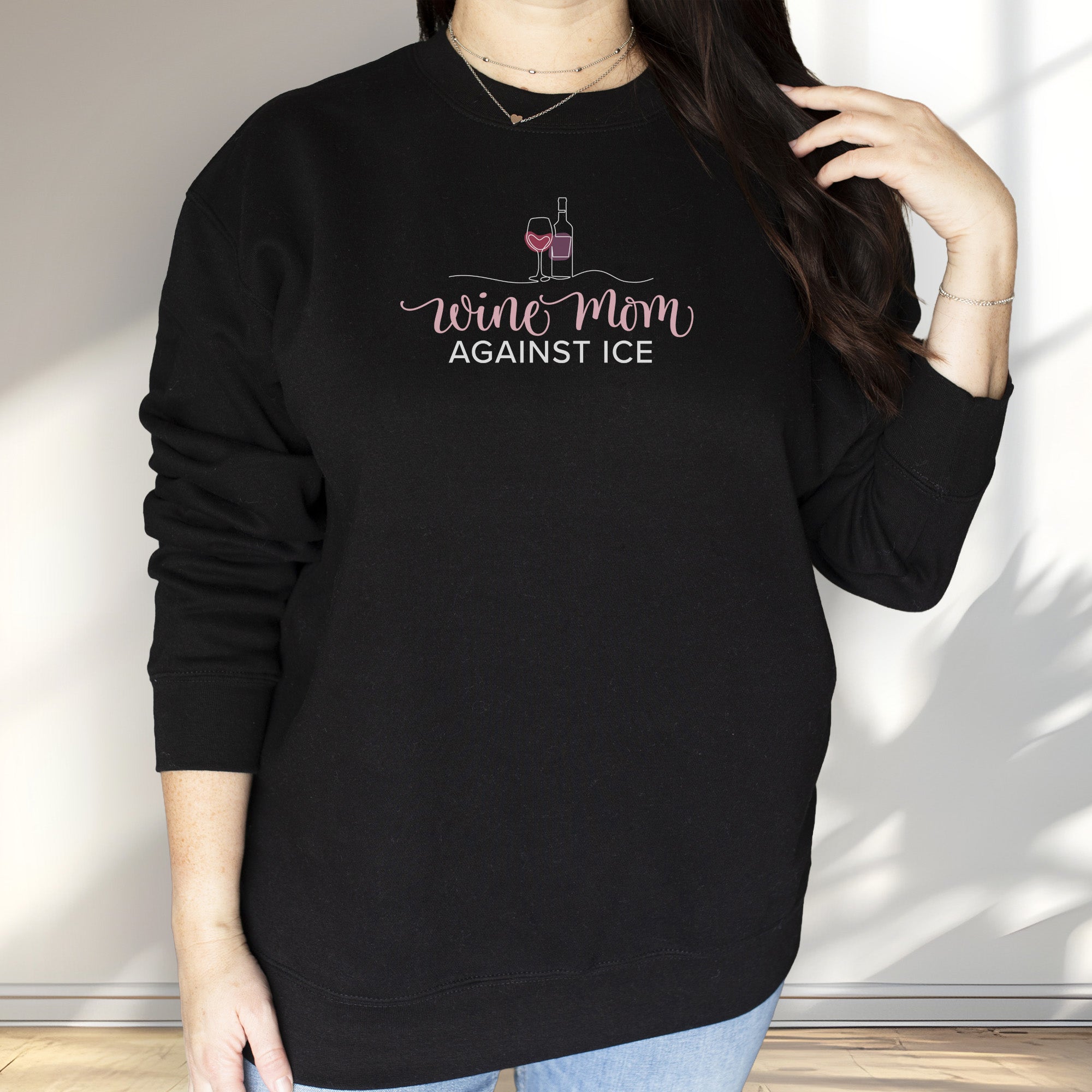 Female model wearing black sweatshirt featuring the text “Wine Mom Against ICE” with a minimalist illustration of a wine bottle and glass above the lettering.