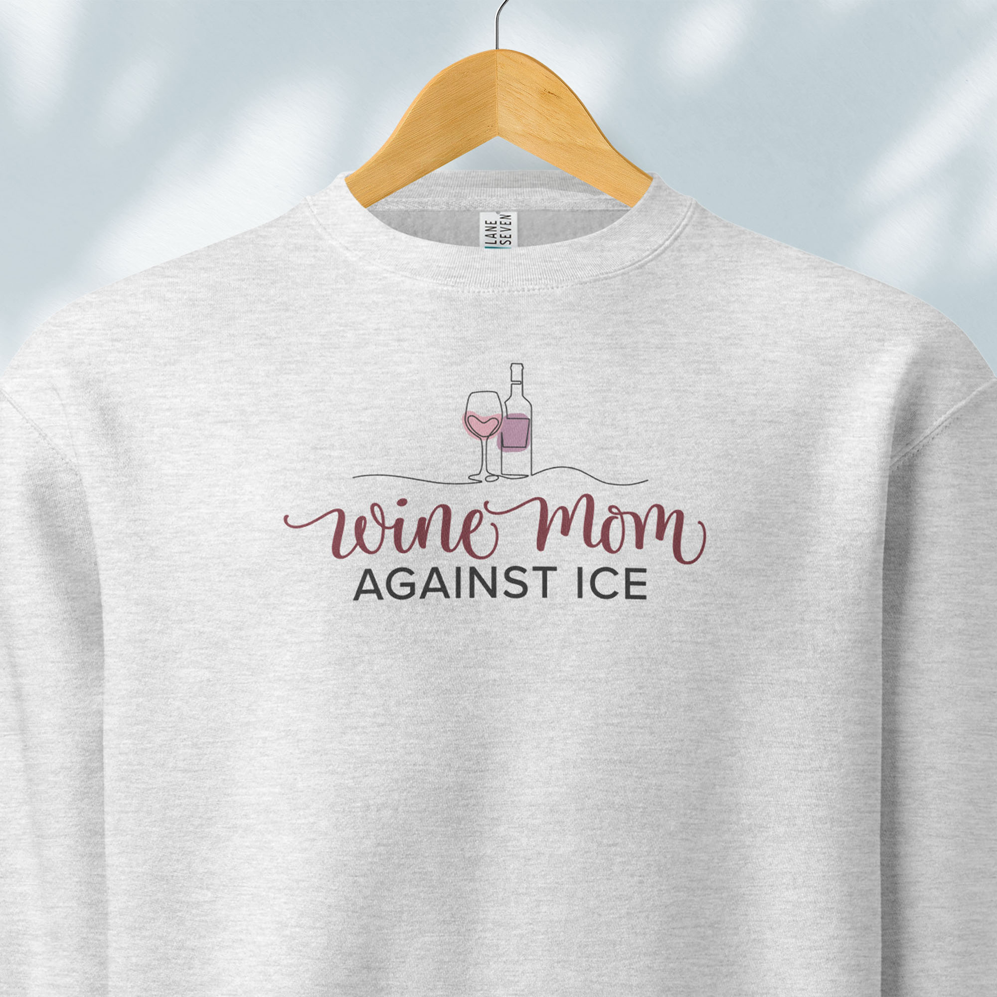 Hanging grey sweatshirt featuring the text “Wine Mom Against ICE” with a minimalist illustration of a wine bottle and glass above the lettering.