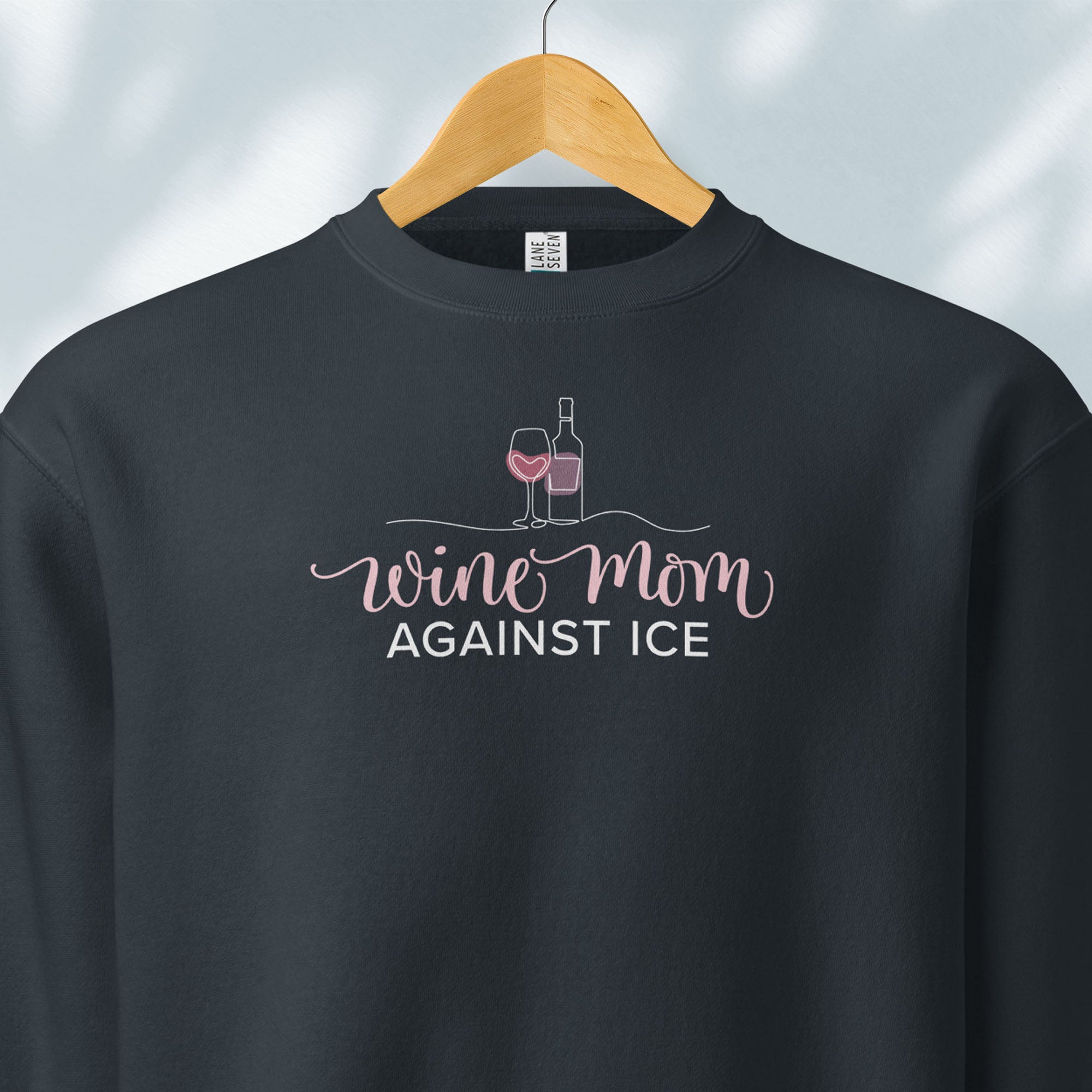 Hanging navy sweatshirt featuring the text “Wine Mom Against ICE” with a minimalist illustration of a wine bottle and glass above the lettering.