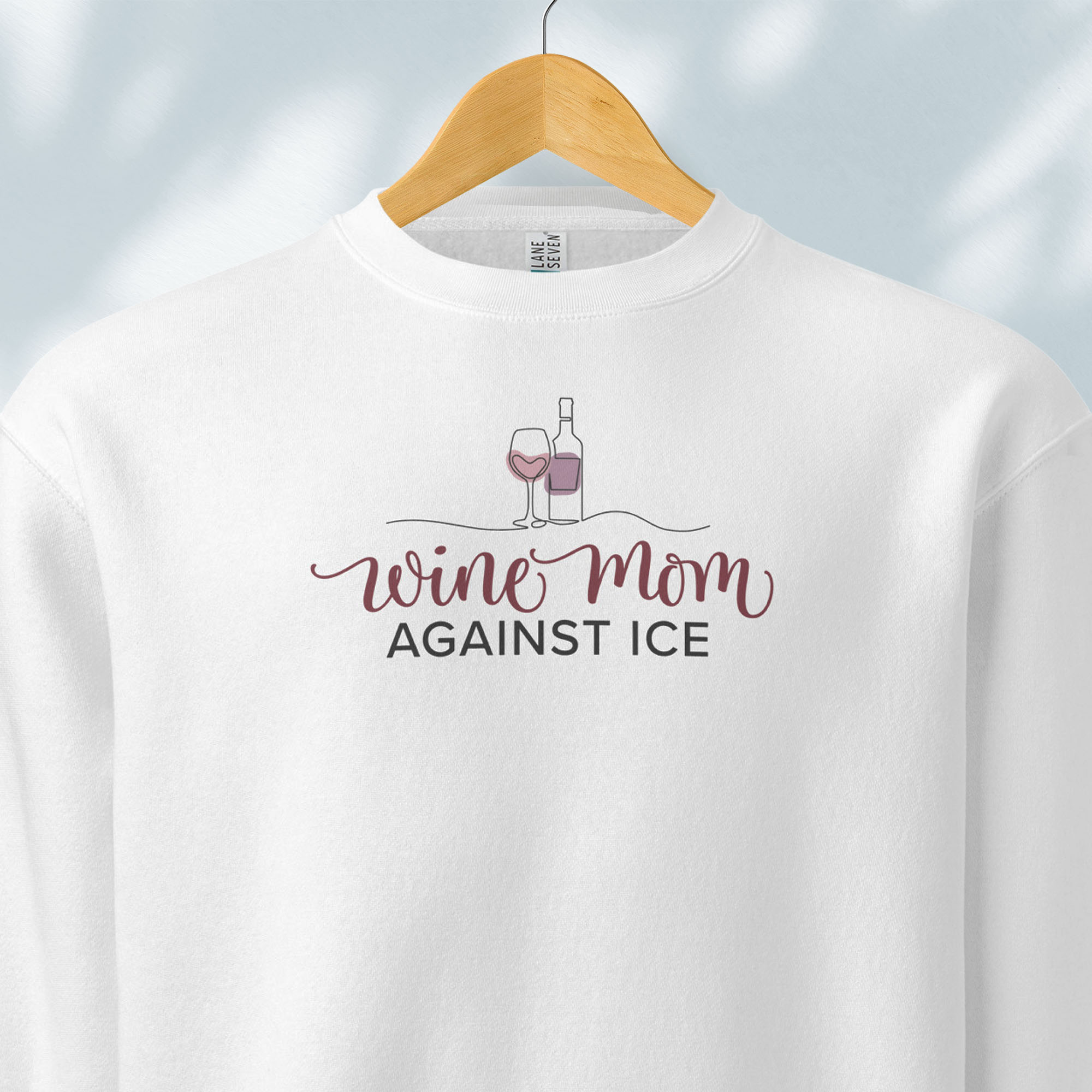 Hanging white sweatshirt featuring the text “Wine Mom Against ICE” with a minimalist illustration of a wine bottle and glass above the lettering.