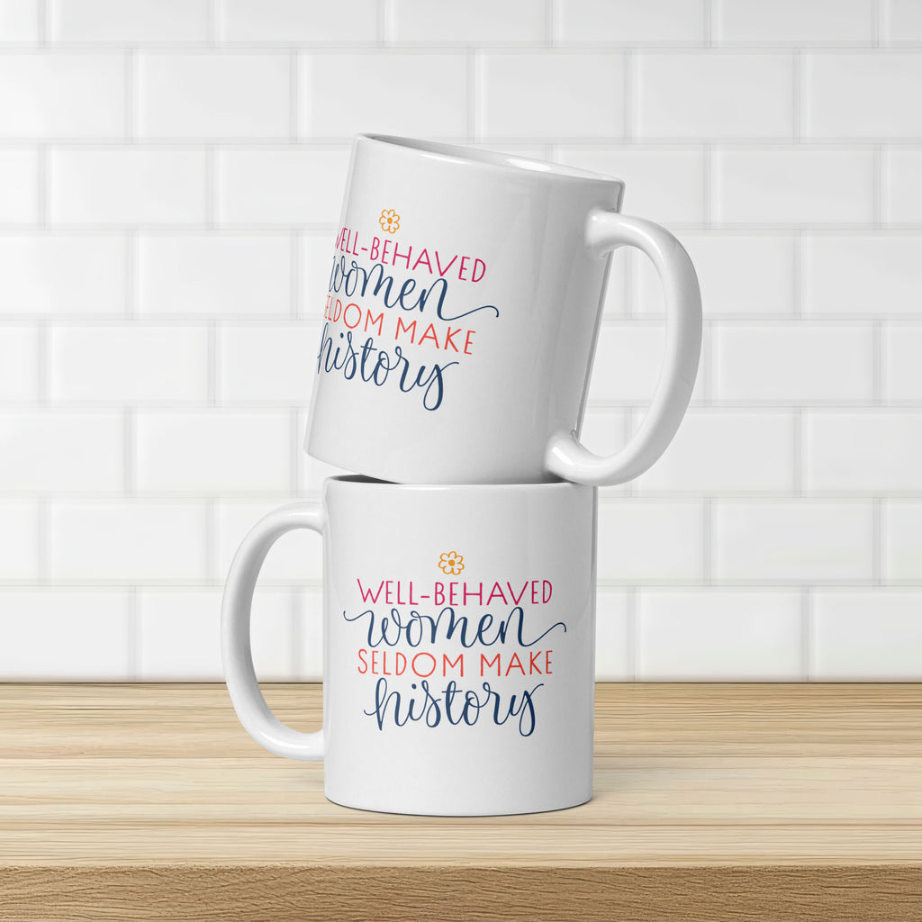 Two white mugs with motivational quotes that are the same on both side "well-behaved women seldom make history" on a wooden surface and tiled wall background.