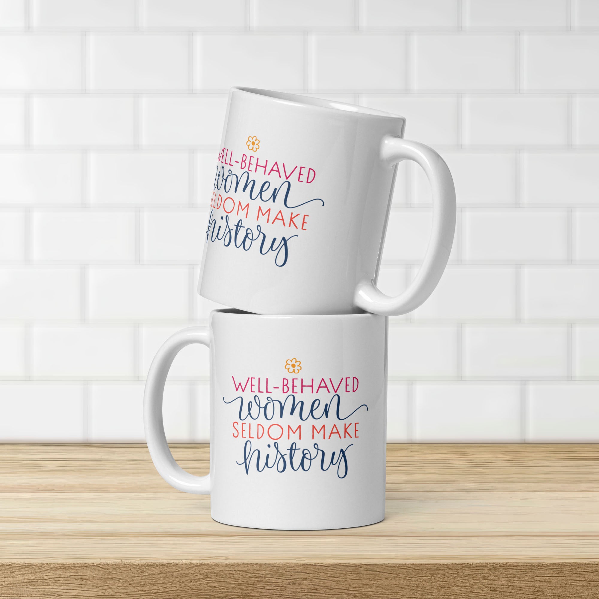 Two white mugs with motivational quotes that are the same on both side "well-behaved women seldom make history" on a wooden surface and tiled wall background.